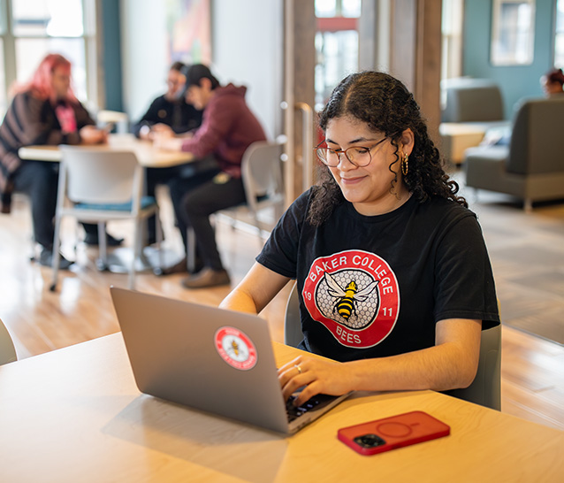Student working on a laptop at a table with students studying in the background.
